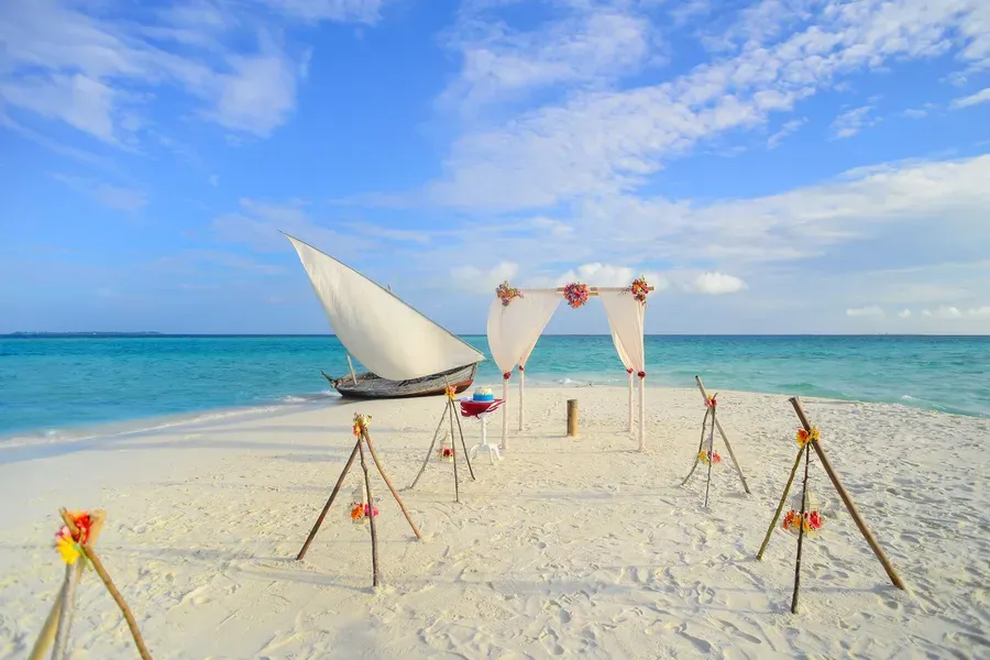 A sailboat anchored offshore beyond a beach wedding arch